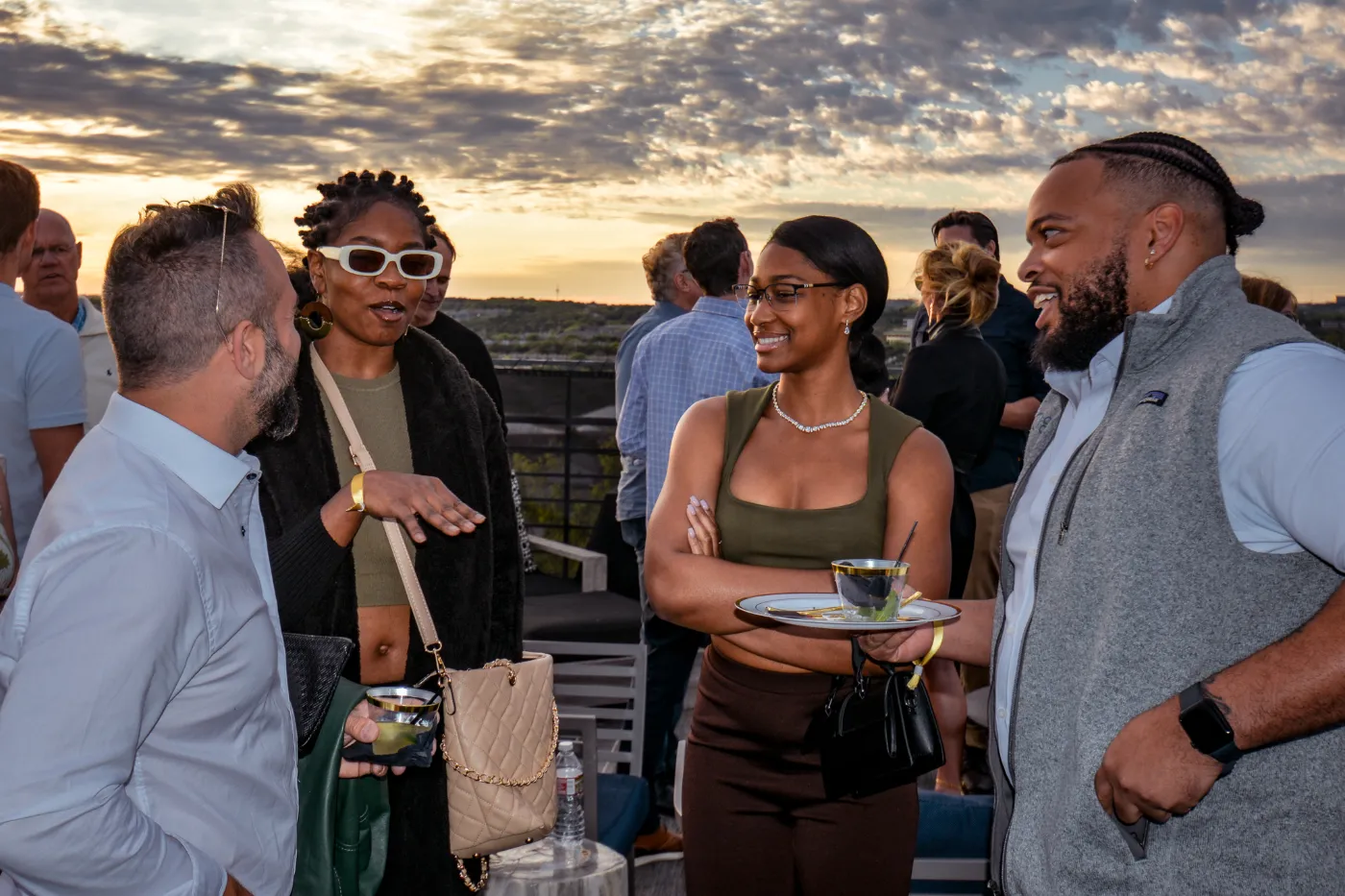 Four people talking on a rooftop terrace at sunset, with more people gathered behind them.
